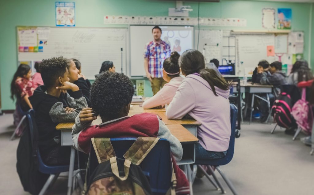 children sitting at desks in classroom with teacher standing at the front of the room